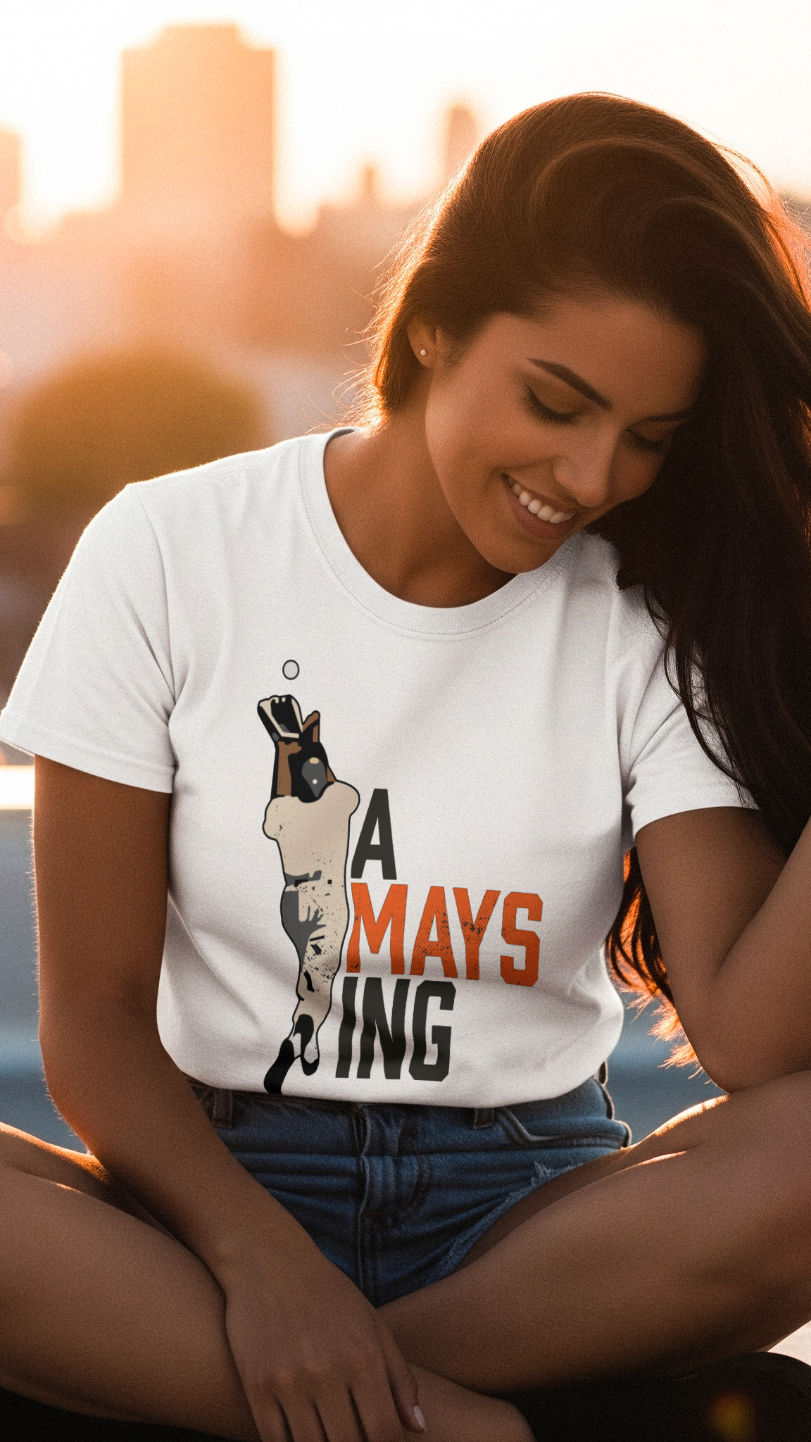 Woman wearing a white t-shirt with a graphic design sitting by a waterfront at sunset.